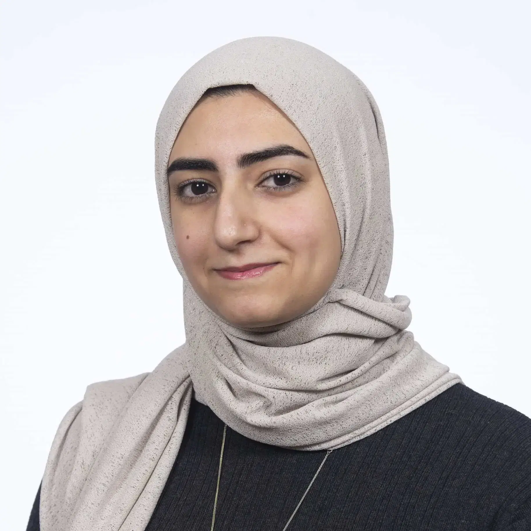 A woman wearing a light beige hijab and a dark jumper looks at the camera with a slight smile against a plain white background.