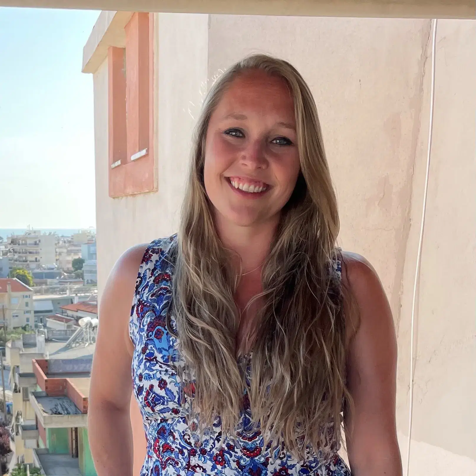 A woman with long blonde hair and a patterned sleeveless top smiles whilst standing on a balcony with a cityscape in the background on a sunny day.