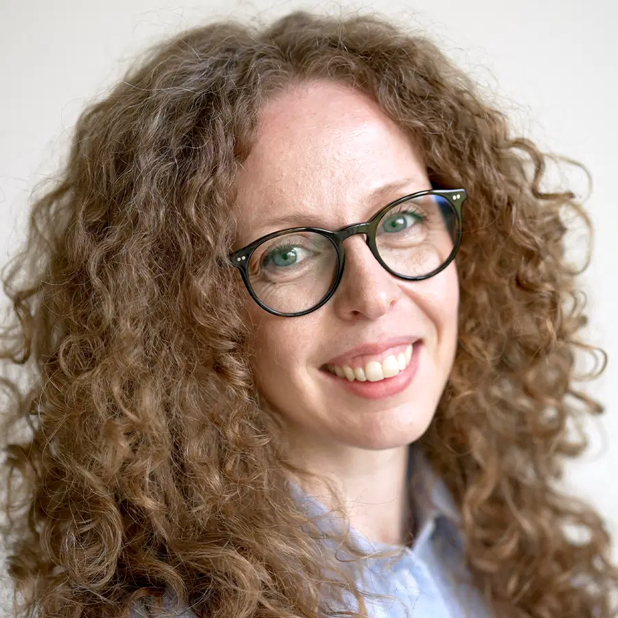 Smiling person with long curly hair and glasses, wearing a light blue shirt, standing in front of a neutral background.