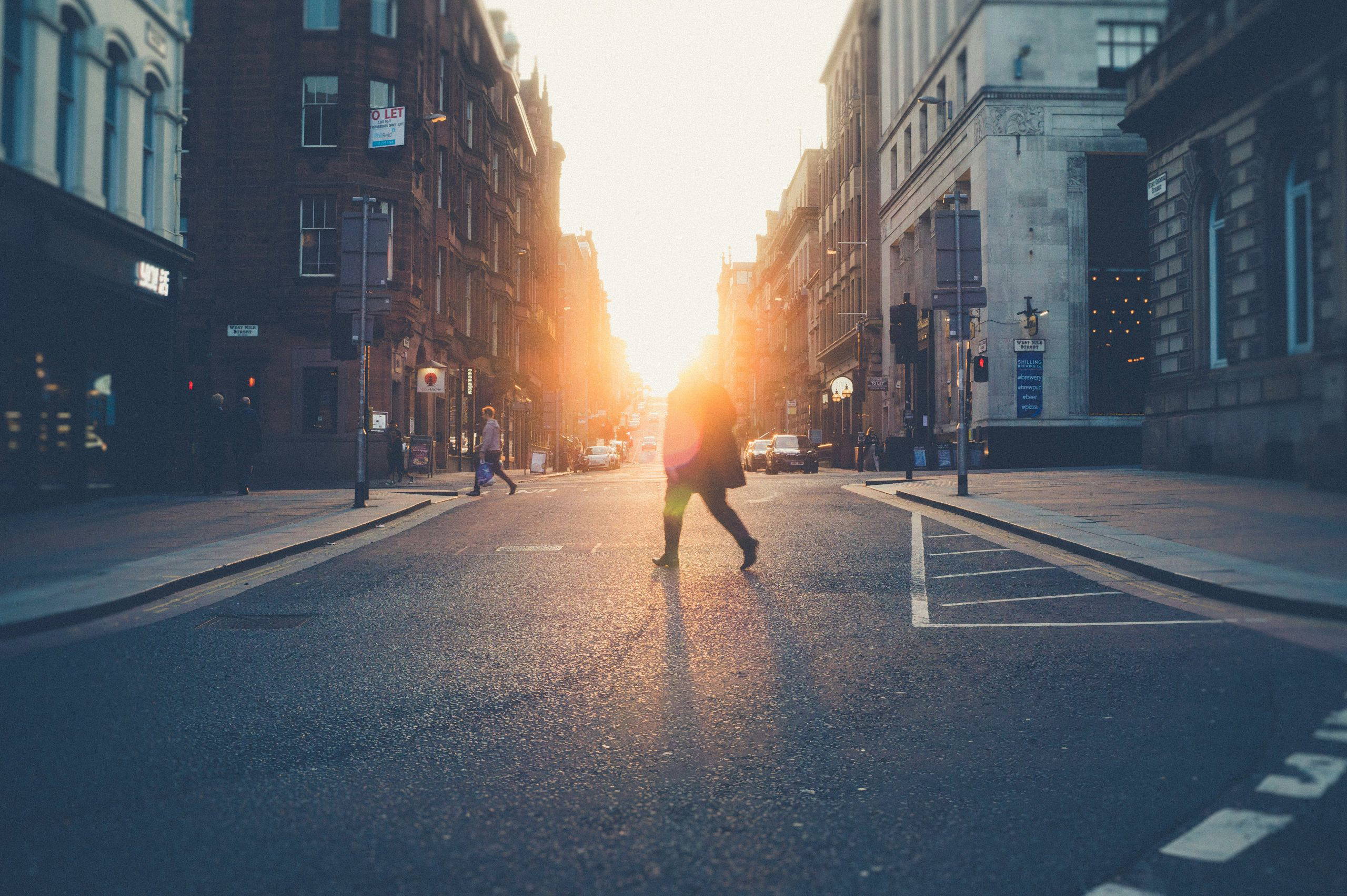 A person walks across a city street at sunset, with tall buildings lining the road on both sides.